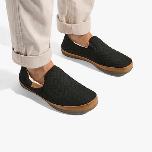 Black quilted slip-on slippers with tan rubber soles and white lining, worn with rolled beige pants.