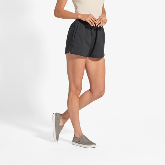 Woman wearing black shorts with taupe grey slip-on shoe with white sole.