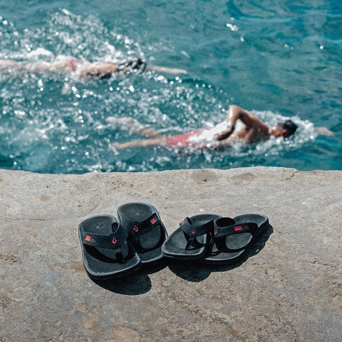 Two pairs of black sandals with red accents sit on concrete by a pool with swimmers in the background.