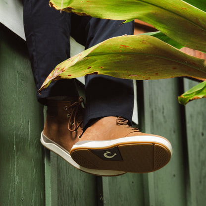 Tan and brown lace-up boot with white sole against green corrugated background and tropical foliage.