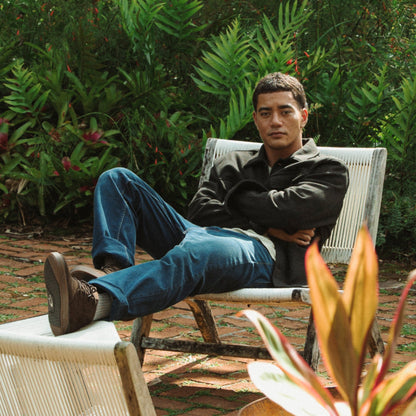Man wearing dark Java jacket and blue jeans seated in white chair against tropical foliage background.