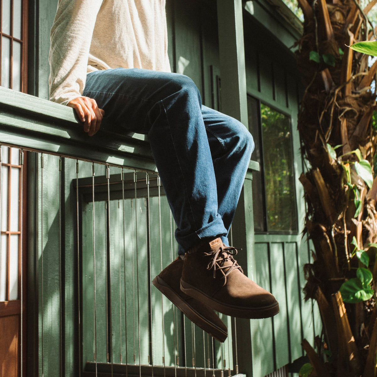 Person wearing dark blue jeans and dark brown suede lace-up boots sitting on a green metal railing.