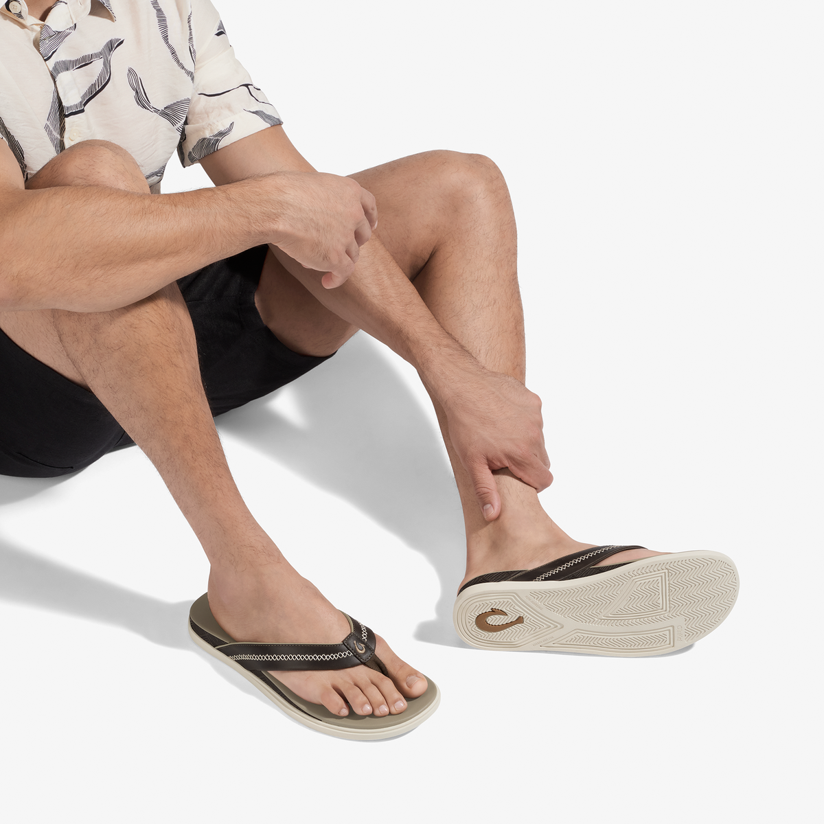 Man wearing cream patterned shirt and black shorts seated while wearing black and cream flip-flops with textured straps.