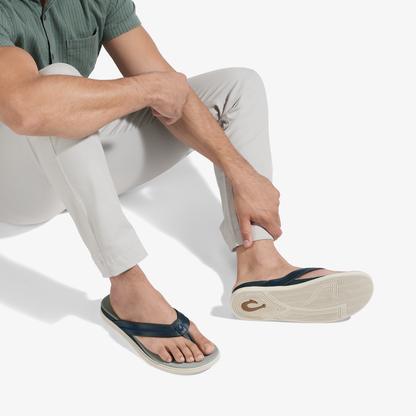 Man wearing pale grey pants and sage green shirt seated, displaying navy and cream thong sandal with visible sole.