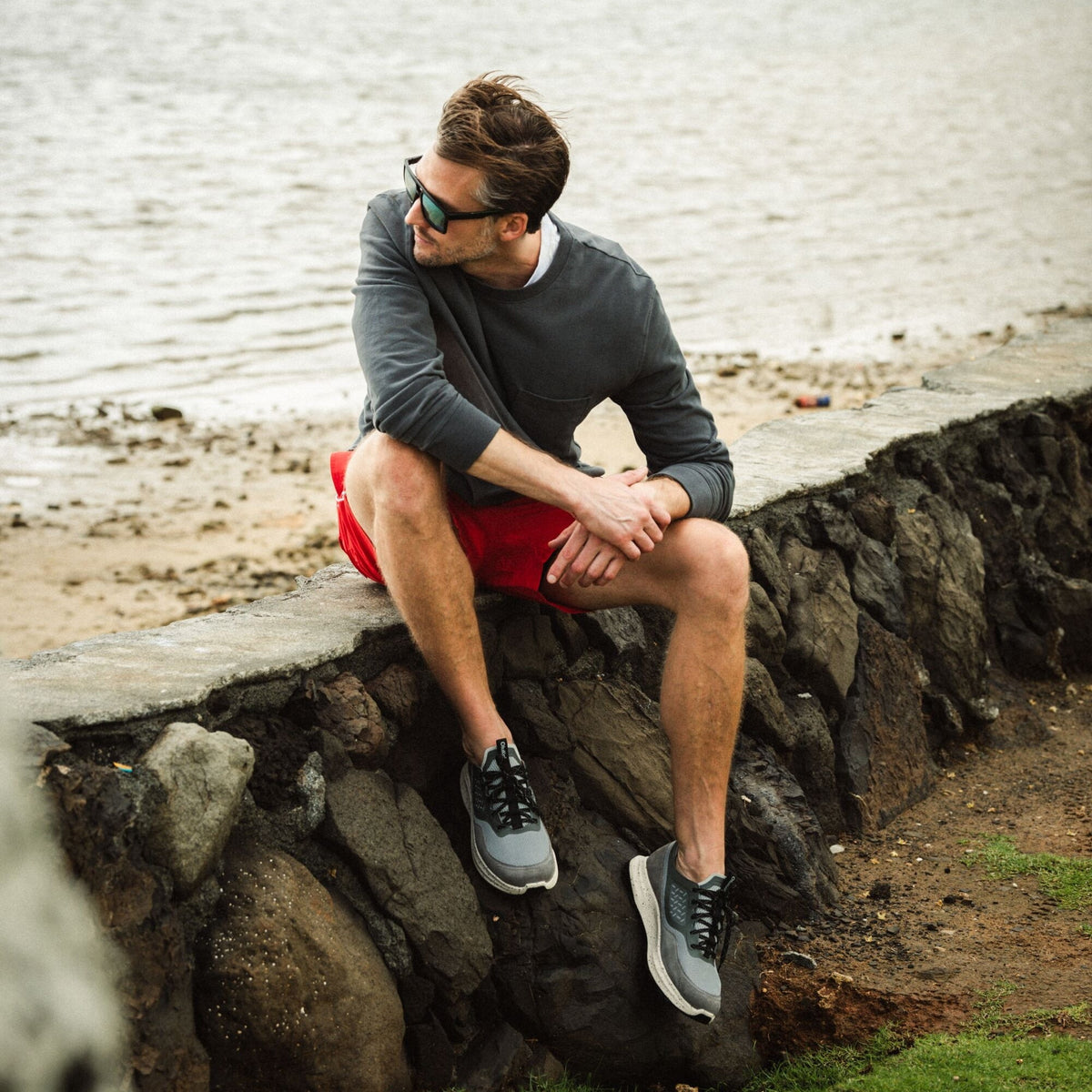 Man wearing grey long-sleeve shirt, red shorts, and grey and black sneakers, sitting on a rock by the water.