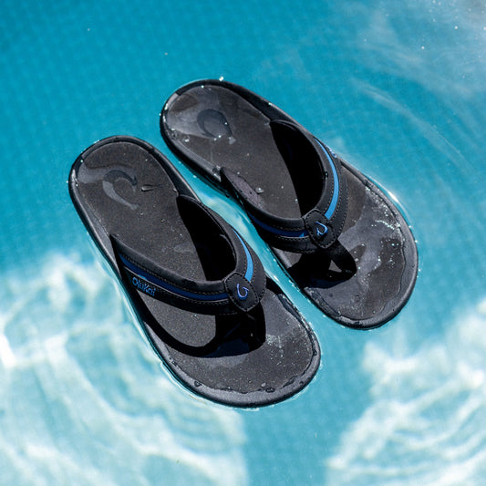 Pair of black and slate blue thong sandals with textured soles, viewed from above on turquoise water.