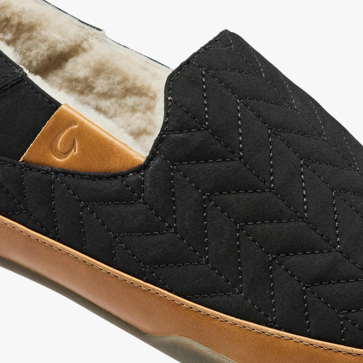 Black quilted slipper with tan leather sole and cream fleece lining, showing curved stitching detail and logo patch.