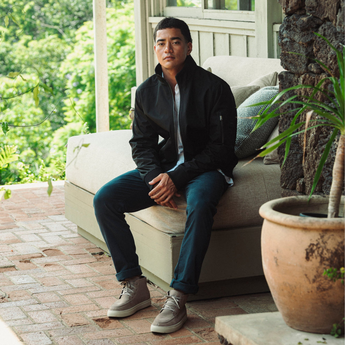 Man wearing taupe grey and tan chukka boots with dark jacket and blue jeans, seated outdoors.