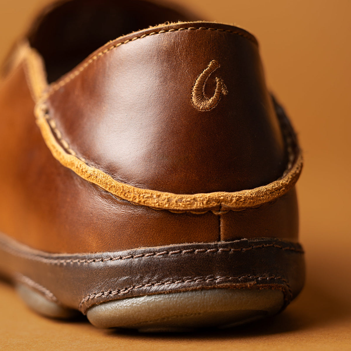 Brown leather slip-on shoe with gold-tone braided trim and embroidered logo detail on heel.