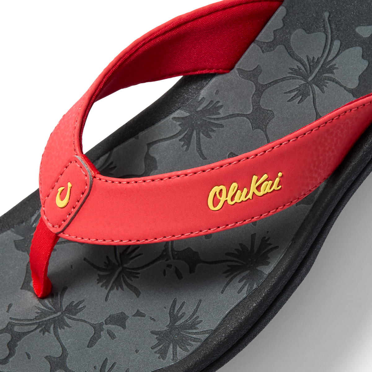 Red flip-flop with yellow "OluKai" script on strap and gray acacia leaf pattern on footbed.