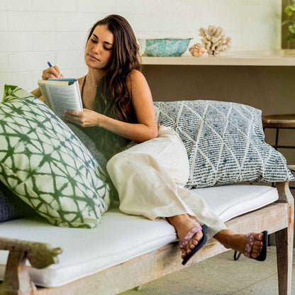 Woman sitting on a light-colored sofa surrounded by decorative pillows in green and blue patterns, holding a notebook.