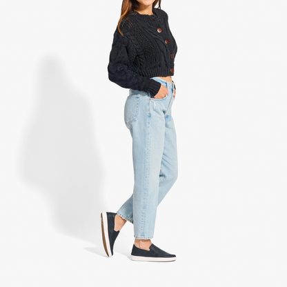 Woman wearing black cable-knit cropped sweater with brown buttons, light blue jeans, and black slip-on sneakers with white so