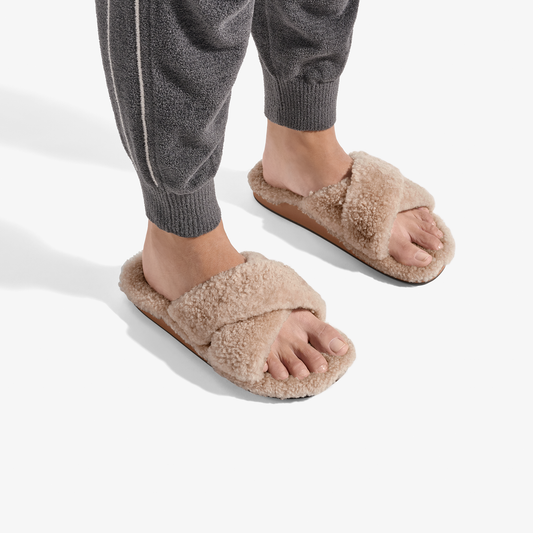 Tan fluffy slide slippers with cushioned footbeds, worn with gray knit pants.