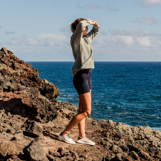 Person wearing a cream-colored long-sleeve shirt and dark shorts standing on rocky coastal terrain overlooking the ocean.