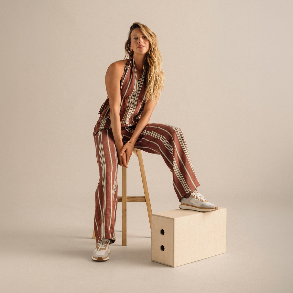 Woman wearing burgundy and cream striped halter jumpsuit with white sneakers, seated on wooden stool.