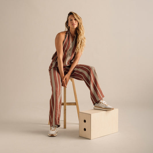 Woman wearing burgundy and cream striped halter jumpsuit with white sneakers, seated on wooden stool.